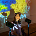 A girl sitting with a standing microphone in front of her and brightly colored letters and words projected on her face